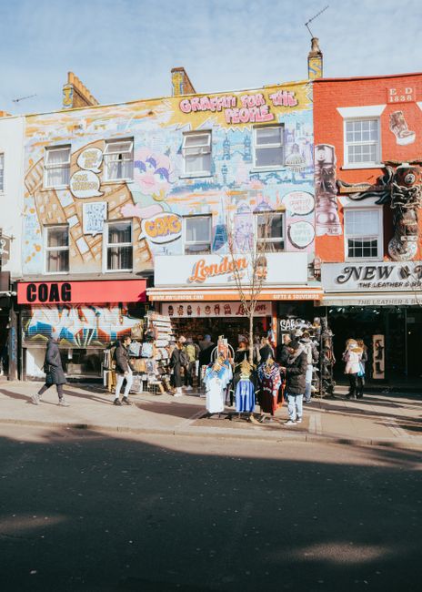 A busy urban street scene under an aged railway bridge with a large, weathered 'Camden Lock' sign painted in yellow on a blue-green metal structure. The ground is paved with cobblestones that appear d